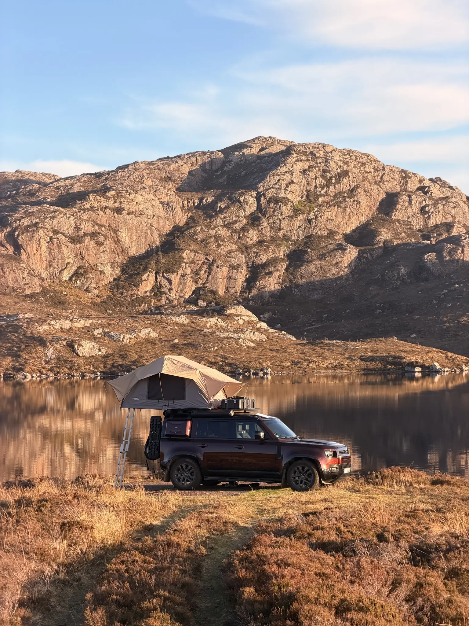Defender 110 in Sedona Red with the rooftop tent open beside a calm loch below a rocky hill